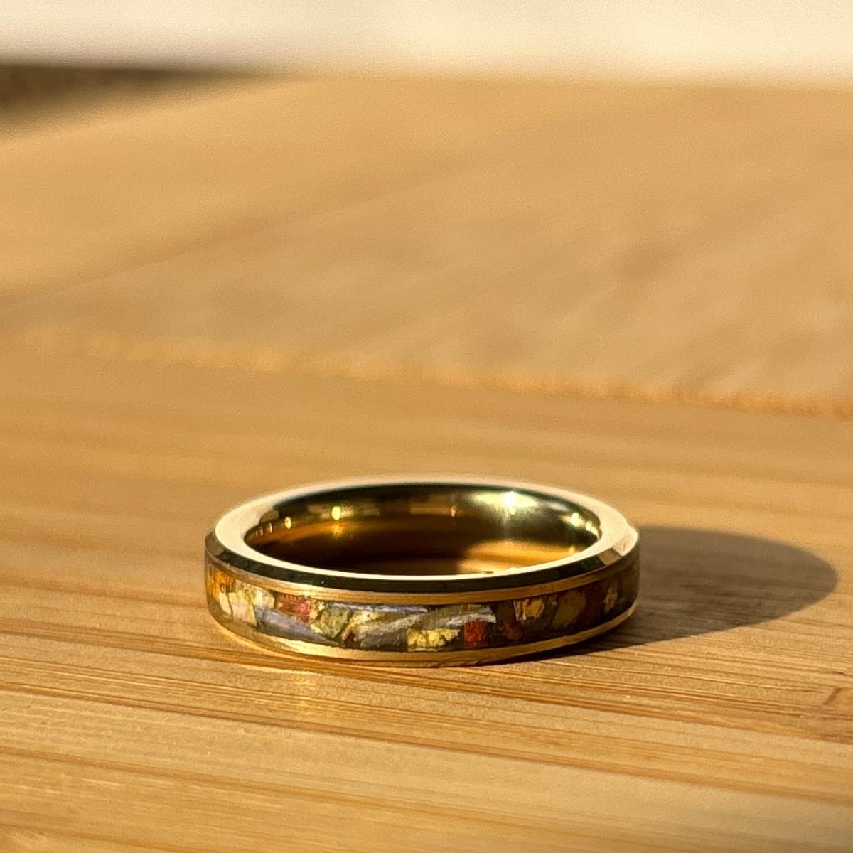 A gold band ring with a colorful inlay of dried leaves, resting on a wooden surface under warm sunlight.
