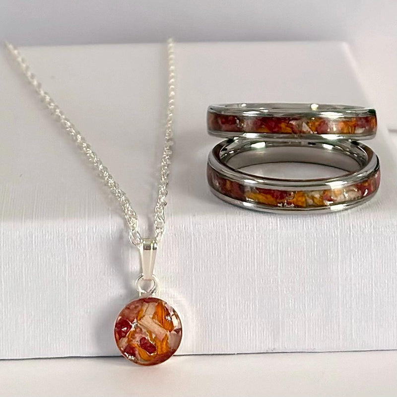 A jewelry set featuring a silver chain necklace with a round pendant and two matching silver rings, all with a warm orange and brown inlay pattern, displayed on a white box.