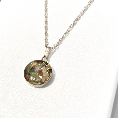 A silver chain necklace with a round pendant containing embedded natural elements like leaves, moss, and small stones, displayed on a textured white surface.