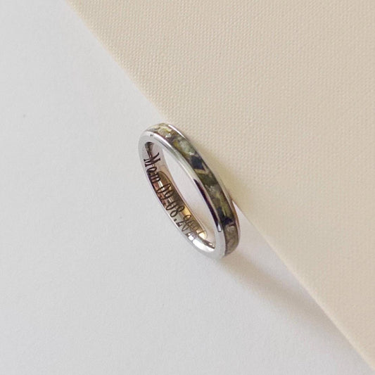 A polished silver band ring filled with dried flowers and resin with engraved script text, resting on a textured cream-colored surface.