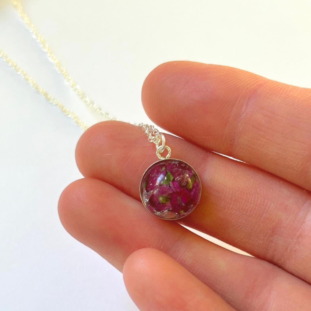 A close-up of a hand holding a small silver pendant necklace with a round domed charm containing pink dried flowers and green leaves, on a white background.
