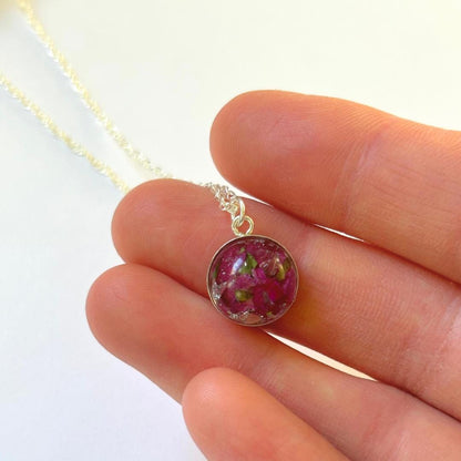 A close-up of a hand holding a small silver pendant necklace with a round domed charm containing pink dried flowers and green leaves, on a white background.