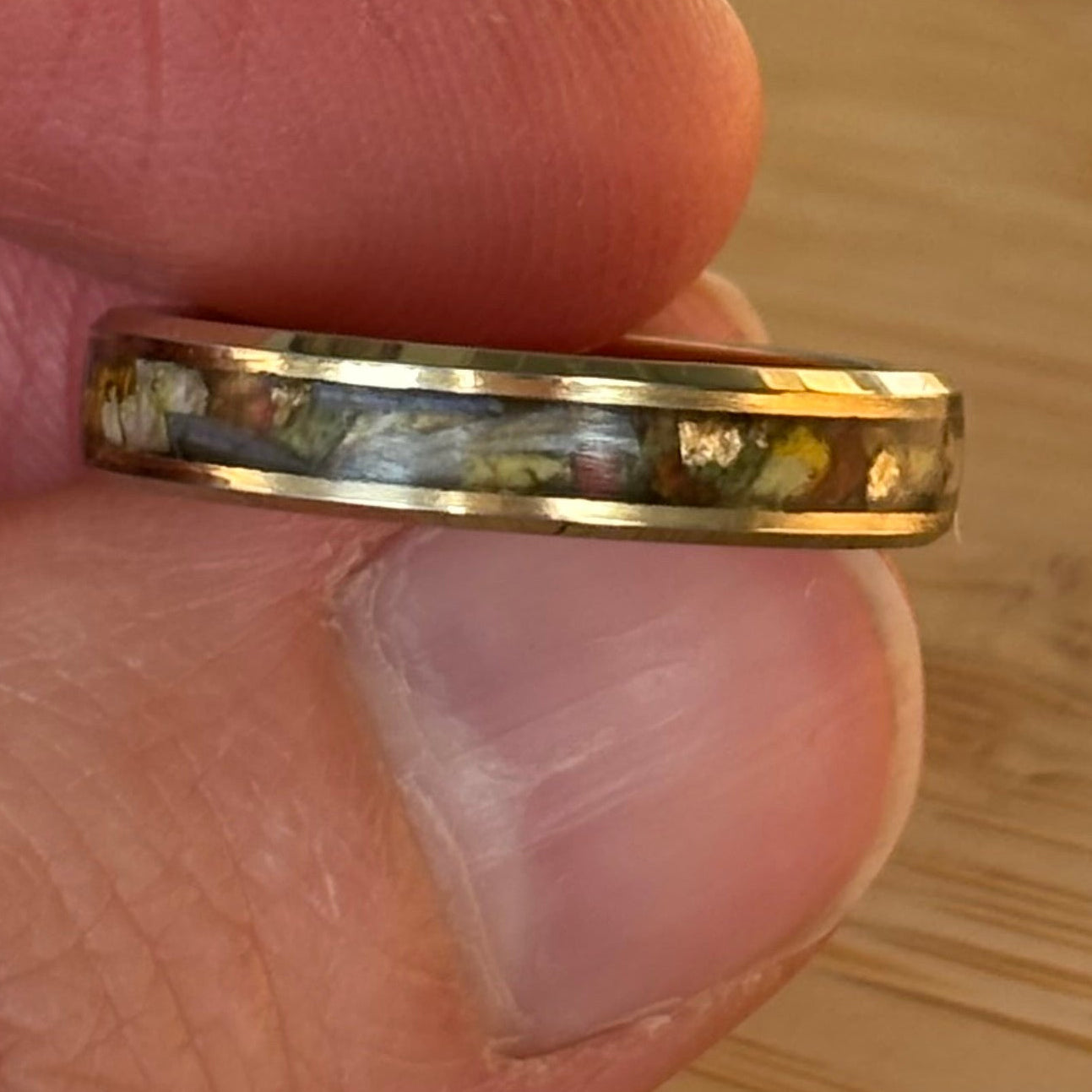 A close-up of a gold band ring held between fingers, featuring a colorful inlay with small gold flecks and a blue-gray center.