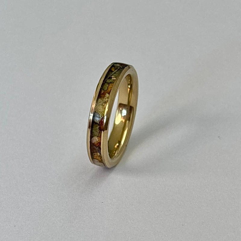 A gold band ring with a colorful inlay of small square tiles, standing upright on a light gray background.