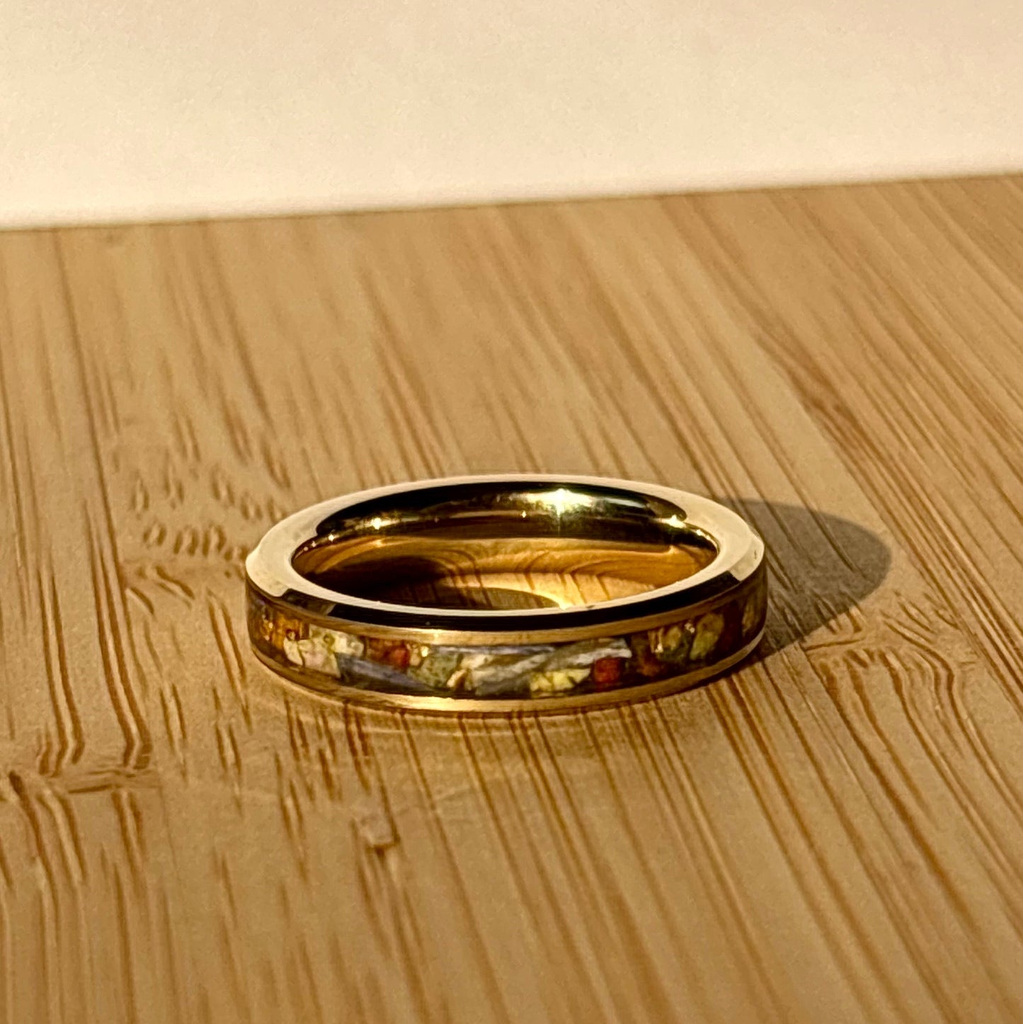 A gold band ring with a colorful inlay of small pieces, resting on a light wood grain surface.