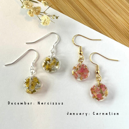 Two pairs of dangle earrings displayed on a white surface with dried flowers and a bamboo-textured background. One pair has silver fishhook wires with clear round drops containing yellow dried flowers, and the other has gold fishhook wires with clear round drops containing pink dried flowers.