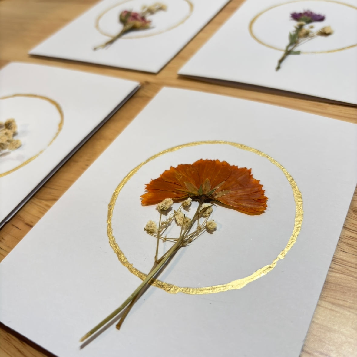 A close-up of a white card featuring a pressed orange flower inside a gold foil circle, placed on a wooden surface with other similar cards partially visible in the background.
