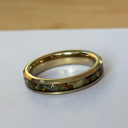 A gold band ring with a colorful inlay of small stones or resin pieces, resting on a white surface with a light wood background.