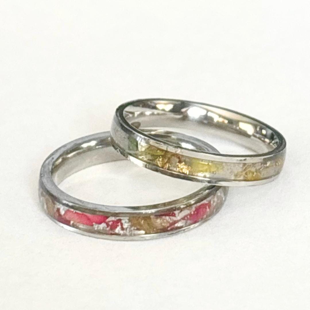Two silver-toned rings with embedded decorative flakes, one featuring pink and white pieces and the other gold and light brown dried flowers, resting on a plain white surface.