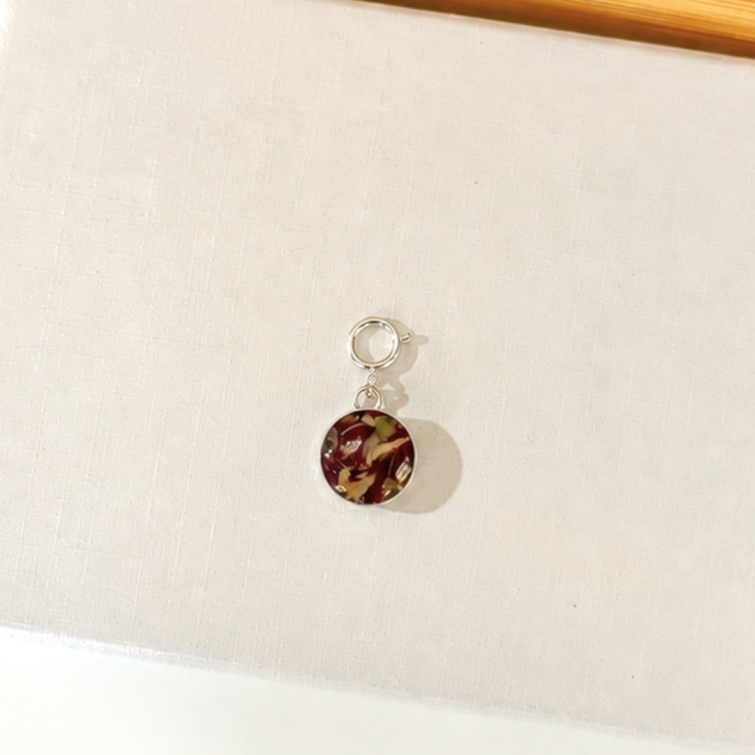 A small silver-toned charm with a circular pendant with dark red, brown, and olive green dried flowers, resting on a textured off-white surface.