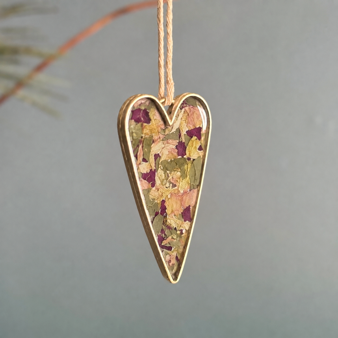 A heart-shaped hanging ornament with a gold-toned metal border and a dried flower inlay, suspended by a thin string against a soft gray background.