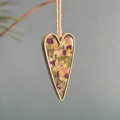A heart-shaped hanging ornament with a gold-toned metal border and a dried flower inlay, suspended by a thin string against a soft gray background.