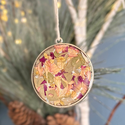 A round silver pendant ornament hanging from a string, filled with dried flowers and leaves in muted pink, purple, green, and gold tones, photographed against a blurred natural background with pine branches and a blue sky.