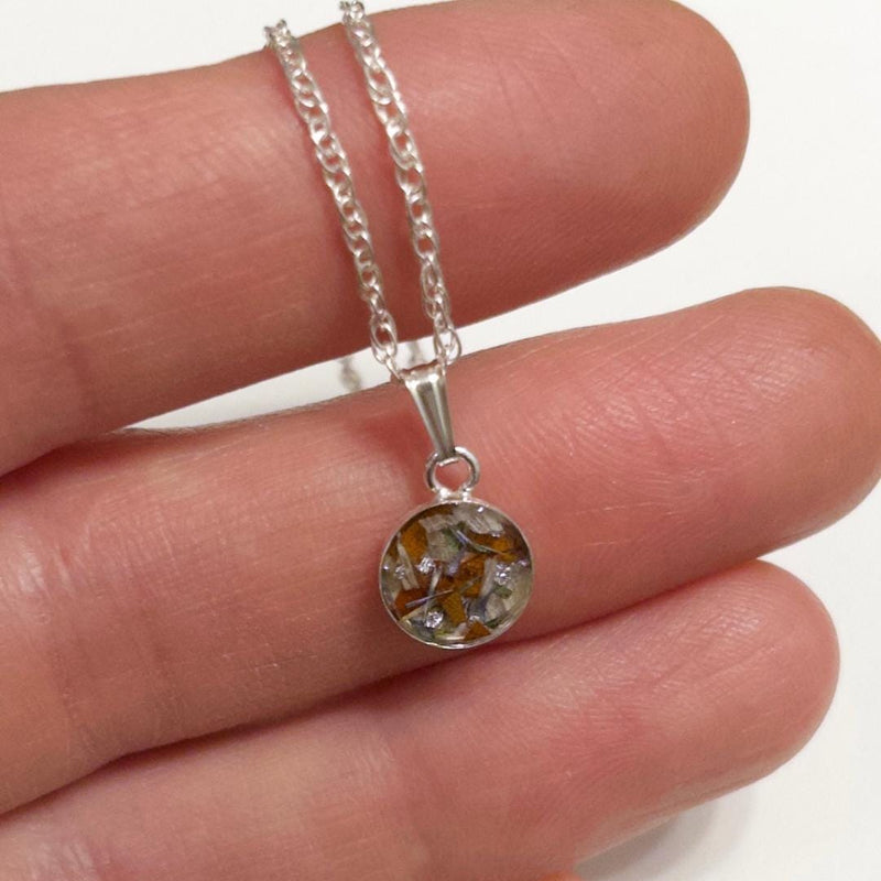 A silver chain necklace with a round pendant containing dried botanical elements, held on a person's fingers against a white background.